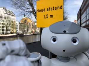 A close-up of the head of a NAO robot, holding out its hand towards the camera as though to push it away, with a sign in the background saying 'keep your distance' in Dutch.