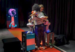 An image of Embrace Angels in action, showing two adult humans hugging one another, while two robotic arms wrap around them in an embrace.